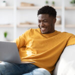 oyful millennial black guy in casual sitting on couch with laptop, surfing on Internet, watching movie or playing game online, looking at notebook screen and smiling, home interior, copy space