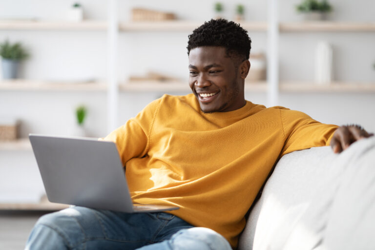 oyful millennial black guy in casual sitting on couch with laptop, surfing on Internet, watching movie or playing game online, looking at notebook screen and smiling, home interior, copy space